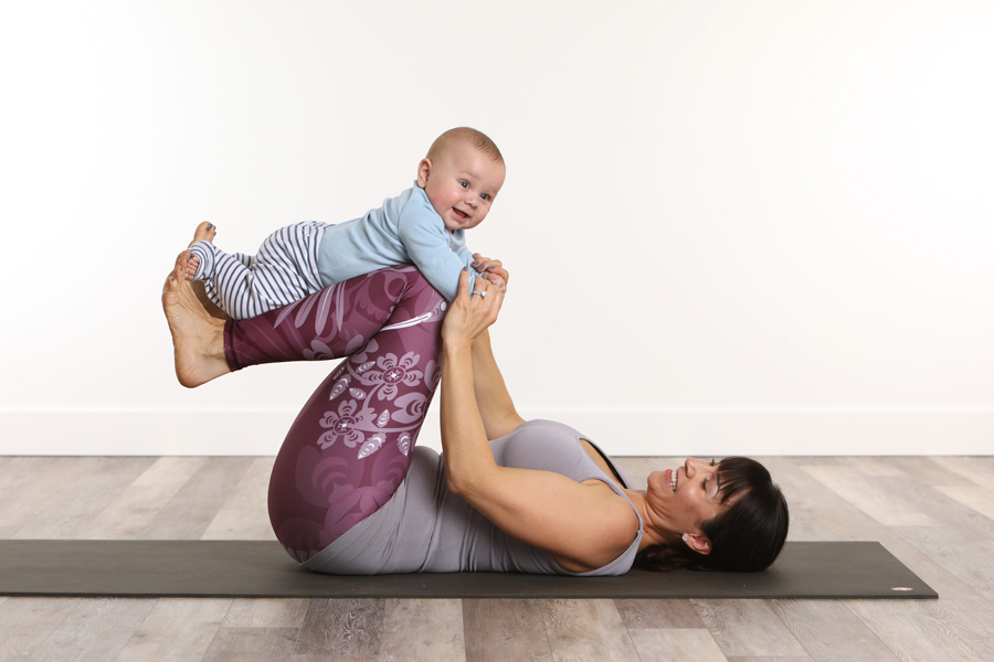 lifestyle photo of mother in yoga pants holding baby on legs while lying on a black yoga mat