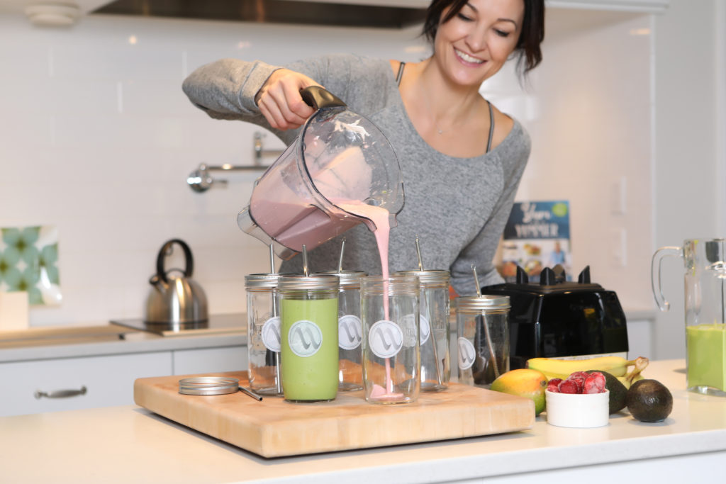 Lifestyle photo of woman in grey top pouring smoothy from blender into mason jars in a kitchen