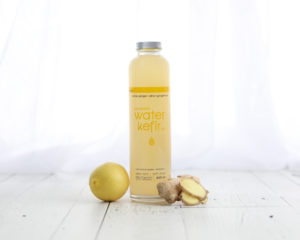 Mini lifestyle photo of lemon water bottle and ginger on white wood in front of white curtains